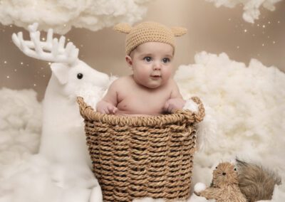 Photographie de bébé en studio, assis dans un panier en osier avec un bonnet tricoté, mise en scène hivernale avec décor féerique et ambiance douce, réalisée par un photographe professionnel spécialisé dans les séances nouveau-né.