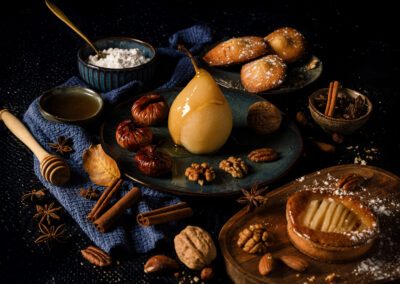 Photographie culinaire d’une poire pochée au miel et d’une tartelette aux poires, dans une ambiance automnale chaleureuse avec fruits secs, épices et douce lumière, réalisée par un photographe professionnel spécialisé dans la photo culinaire.