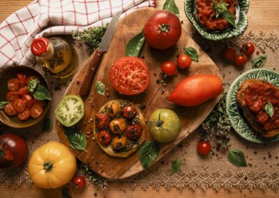 Photographie culinaire mettant en scène des tomates de différentes variétés avec basilic, huile d’olive et tartines, dans une ambiance rustique et lumineuse évoquant la cuisine méditerranéenne. Réalisée par un photographe culinaire professionnel.