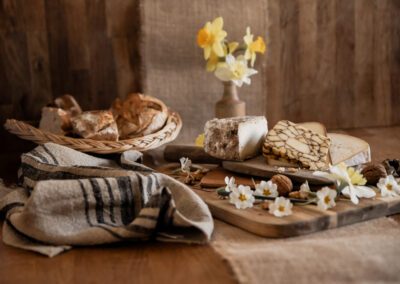 Photographie culinaire mettant en scène un plateau de fromages artisanaux et de pain frais, décoré de fleurs et de noix, dans une ambiance rustique et naturelle. Réalisée par un photographe culinaire professionnel spécialisé dans les produits du terroir.