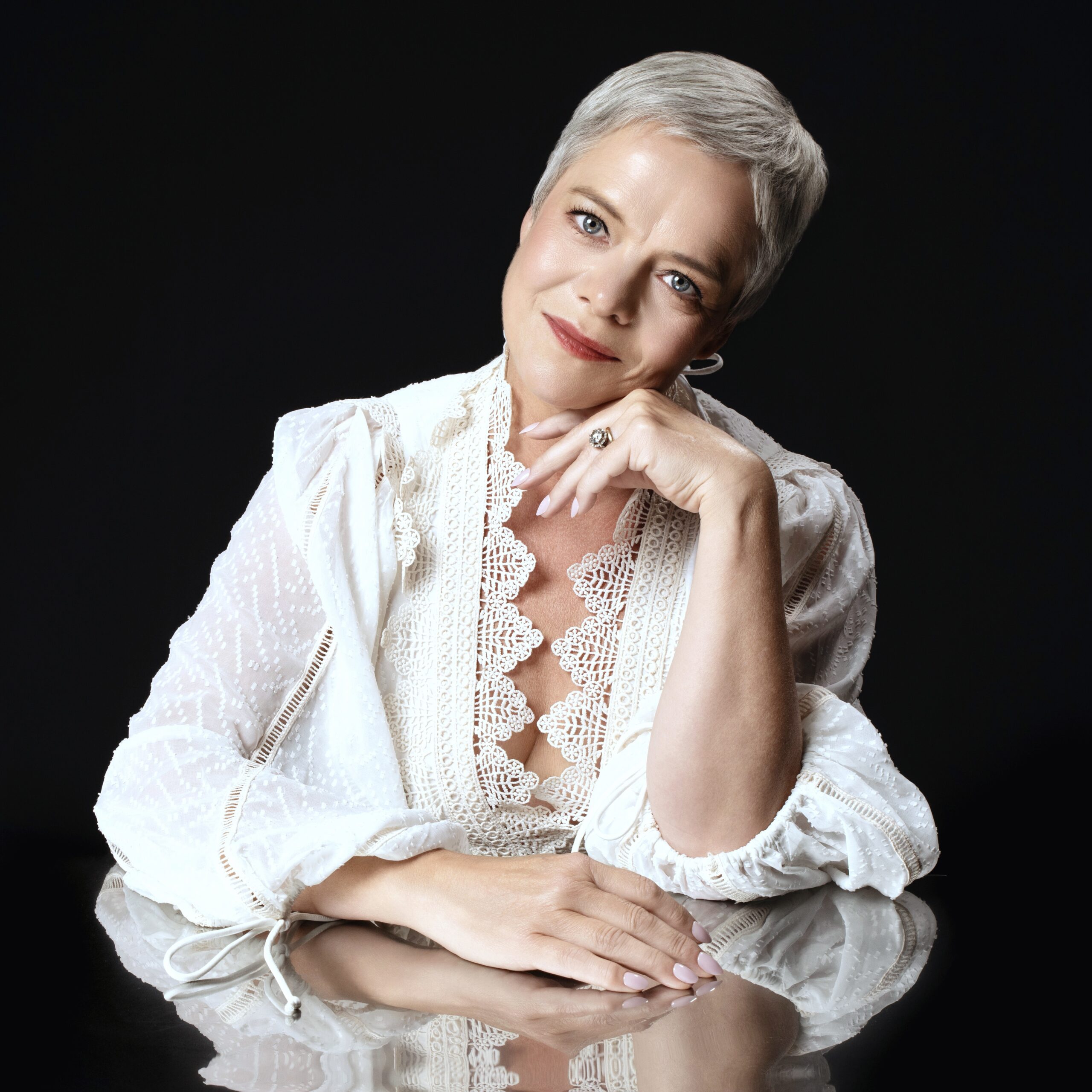Femme aux cheveux gris courts, vêtue d’une blouse blanche en dentelle, posant avec douceur et élégance sur fond noir. Portrait réalisé en studio, lumière douce et regard bienveillant.