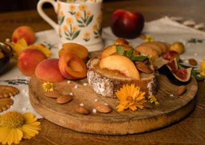 Photographie culinaire d’une tartine aux fruits d’été — pêches, abricots et amandes — mise en scène sur une table en bois avec fleurs, biscuits et vaisselle, réalisée par un photographe professionnel spécialisé dans les ambiances naturelles et gourmandes.
