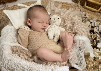 Photographie de nouveau-né réalisée en studio, bébé endormi dans un décor naturel aux tons beiges, accompagné d’une peluche en crochet et de fleurs séchées, capturée par un photographe professionnel spécialisé dans les séances bébé.