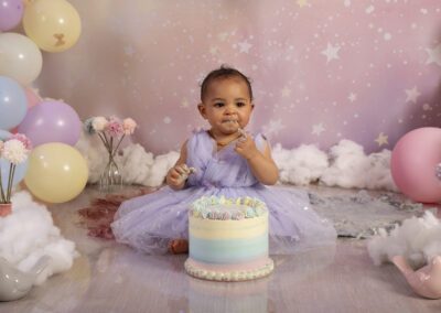 Photographie de séance cake smash en studio, bébé en robe violette dégustant son gâteau d’anniversaire dans un décor pastel avec ballons, fleurs et nuages, réalisée par un photographe professionnel spécialisé dans les photos de bébé.