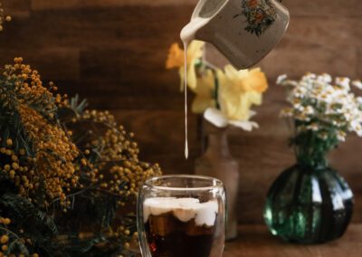Photographie culinaire capturant un geste de lait versé dans un café glacé, mise en scène avec des fleurs printanières et une lumière naturelle. Réalisée par un photographe culinaire professionnel spécialisé dans les compositions poétiques et artisanales.