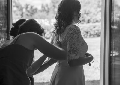 Préparatifs de mariage en noir et blanc : une femme aide la mariée à fermer sa robe en dentelle, près d’une fenêtre, dans une lumière douce et naturelle