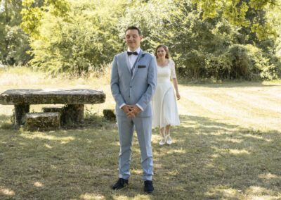 Marié debout en costume clair, les yeux fermés, attendant sa future épouse lors d’un moment de découverte en extérieur, tandis que la mariée s’approche derrière lui dans un cadre naturel et lumineux.