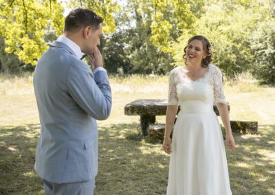 Mariés se faisant face pour la première fois lors de leur découverte en extérieur, le marié ému portant la main à son visage tandis que la mariée sourit, entourés par la nature et une lumière douce.