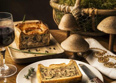 Part de pâté en croûte aux champignons servie sur une assiette blanche, photographiée sur une table en bois. Mise en scène culinaire rustique avec panier, champignons décoratifs et lumière chaude.