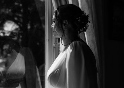 Mariée photographiée en noir et blanc, de profil près d’une fenêtre, regard tourné vers l’extérieur, avec son reflet visible dans la vitre et une lumière douce soulignant ses traits