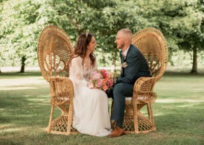 Couple de mariés assis face à face sur des fauteuils en rotin lors d’une cérémonie laïque en extérieur, échangeant un regard complice dans un parc arboré