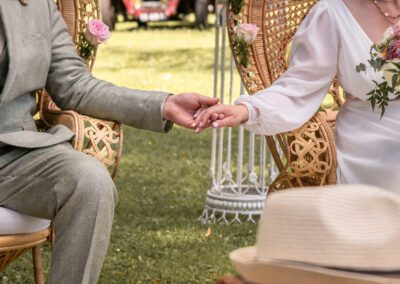 Mariés assis sur des fauteuils en osier lors d’une cérémonie de mariage en plein air, se tenant la main, invités en arrière-plan, ambiance naturelle et intimiste.