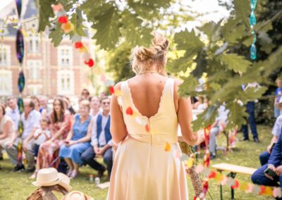 Témoin de mariage vue de dos lors d’une cérémonie en plein air, faisant face aux invités assis sous les arbres, décorations suspendues dans le feuillage, ambiance naturelle et lumineuse.
