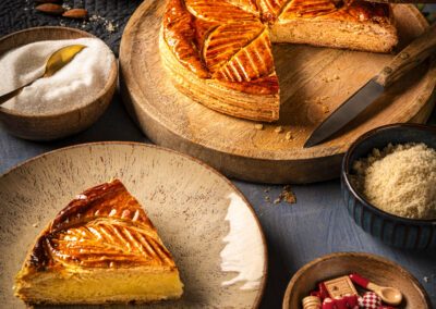 Galette des rois à la frangipane dorée, part découpée et servie sur assiette. Mise en scène culinaire en studio avec couronne, amandes, œufs et lumière chaleureuse.