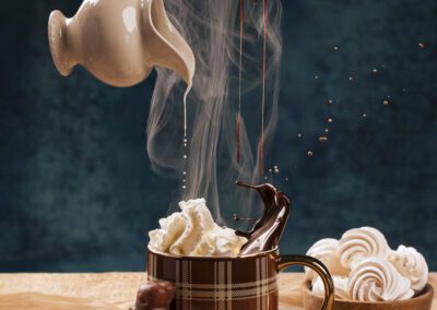 Tasse de chocolat chaud avec crème fouettée, nappée de chocolat fondu et de lait versé en suspension. Mise en scène gourmande en studio, ambiance chaleureuse et hivernale.