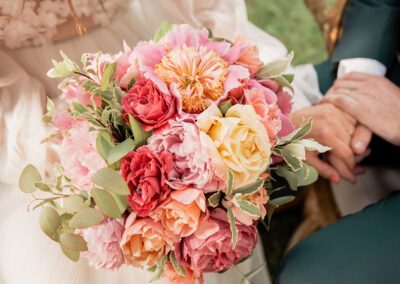 Gros plan sur un bouquet de mariée composé de pivoines et roses aux tons roses, corail et crème, tenu pendant une cérémonie de mariage en extérieur